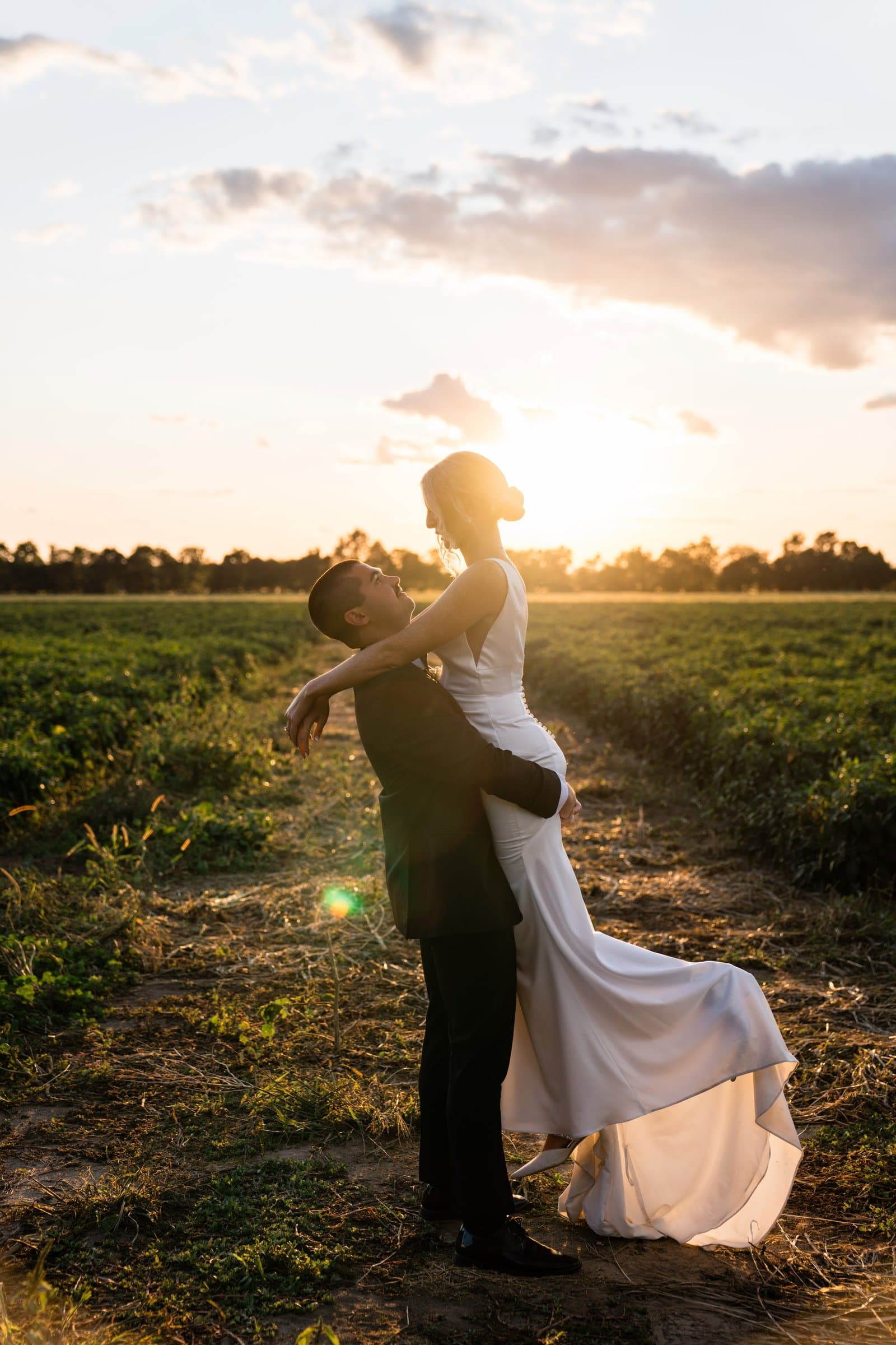 Groom lifting his bride in a farm field as the sun sets behind them