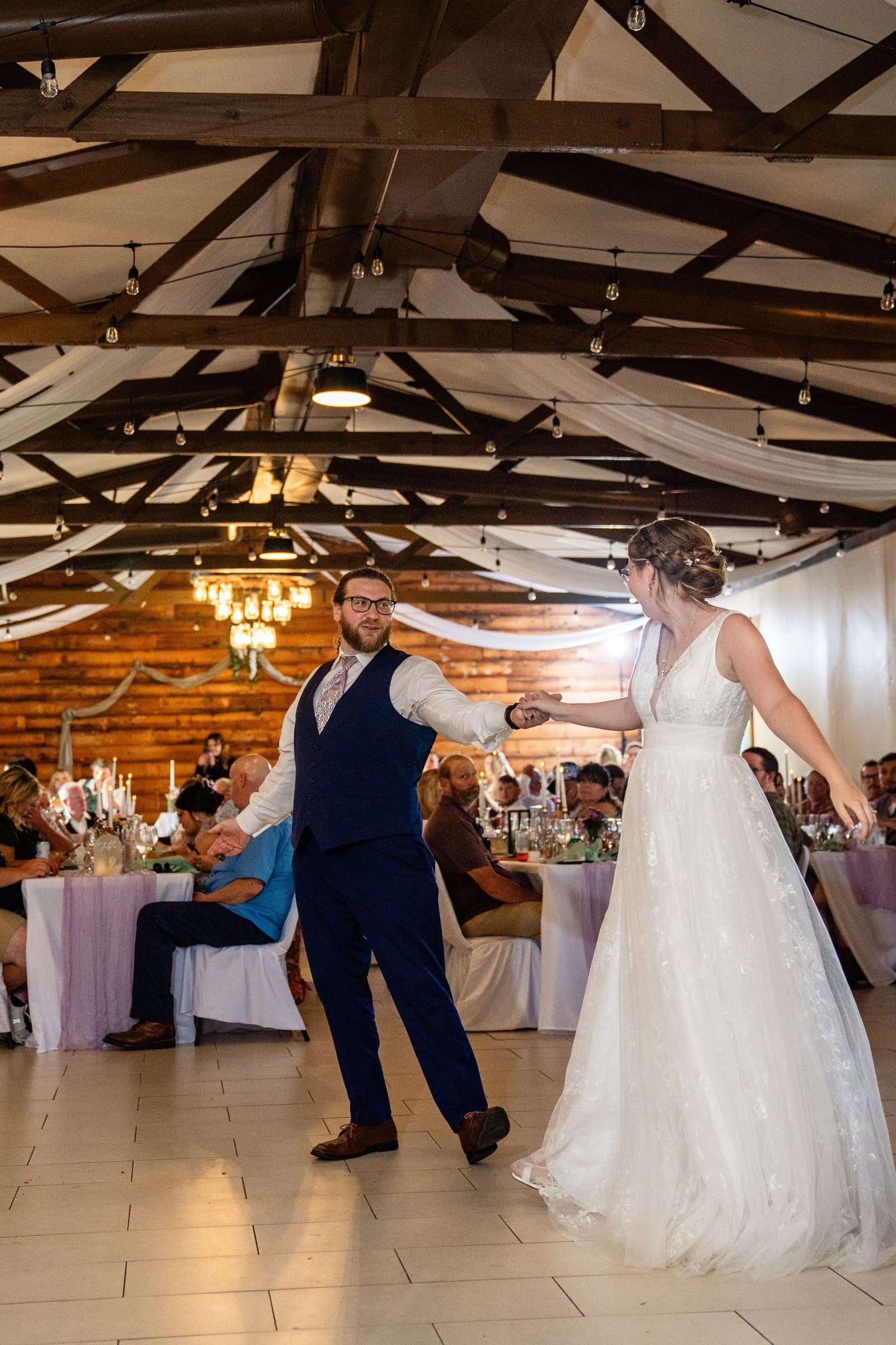Bride and groom dancing in a lit barn reception hall as guests watch from their tables