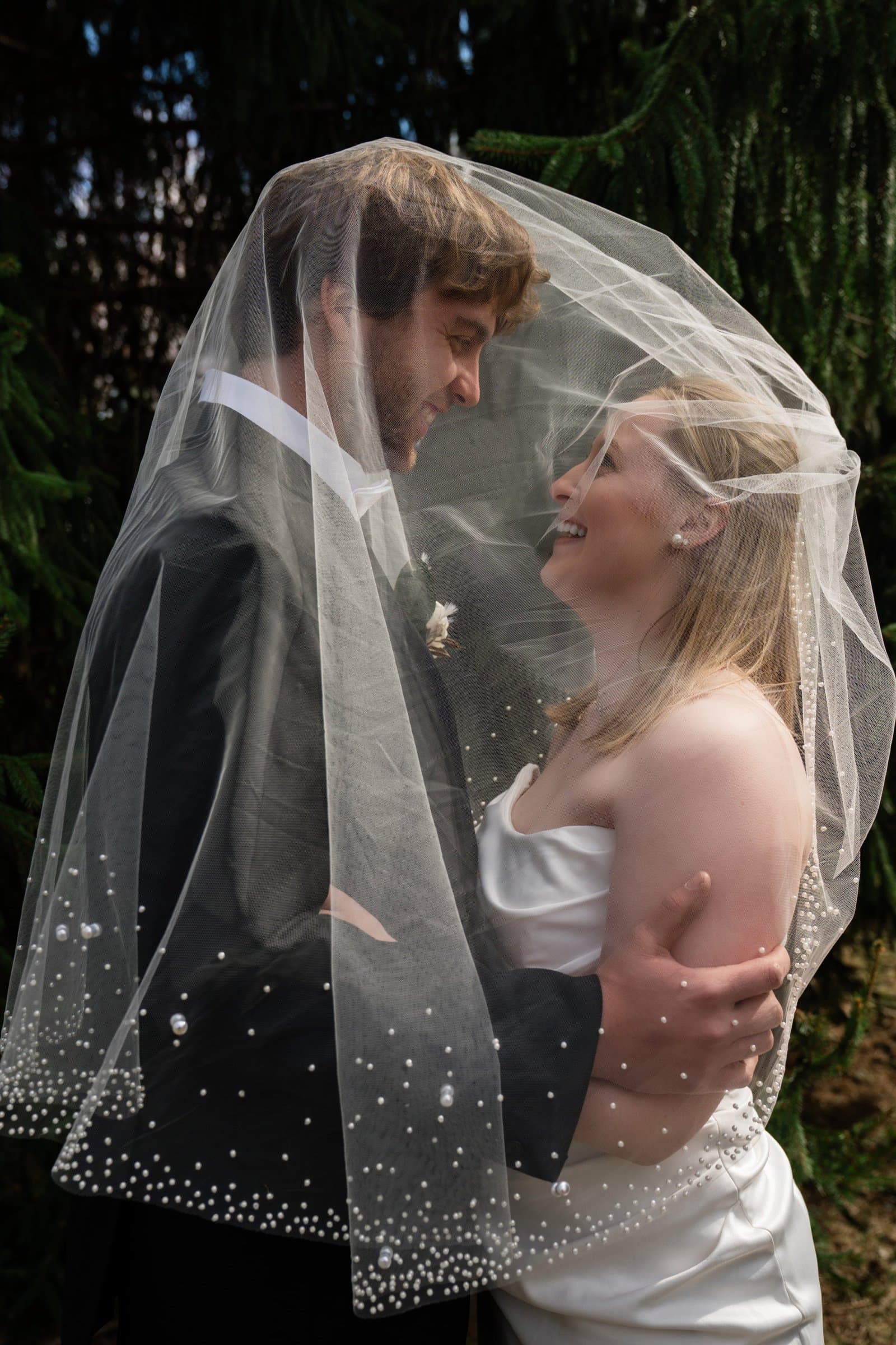 Classic portrait of a bride and groom wrapped in a pearl-edged veil against a dark background