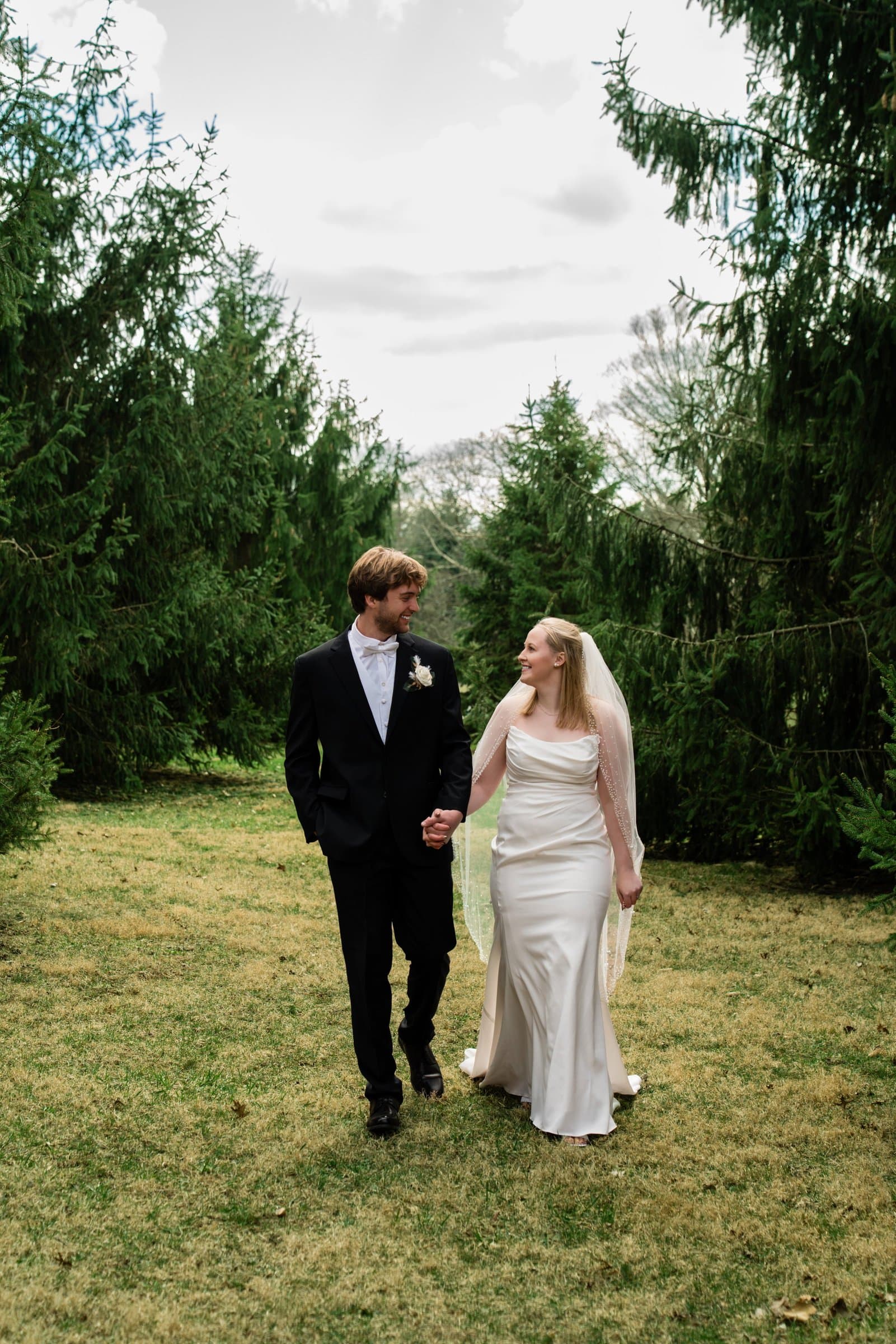 Bride and groom walking hand in hand across a lawn lined with evergreen trees
