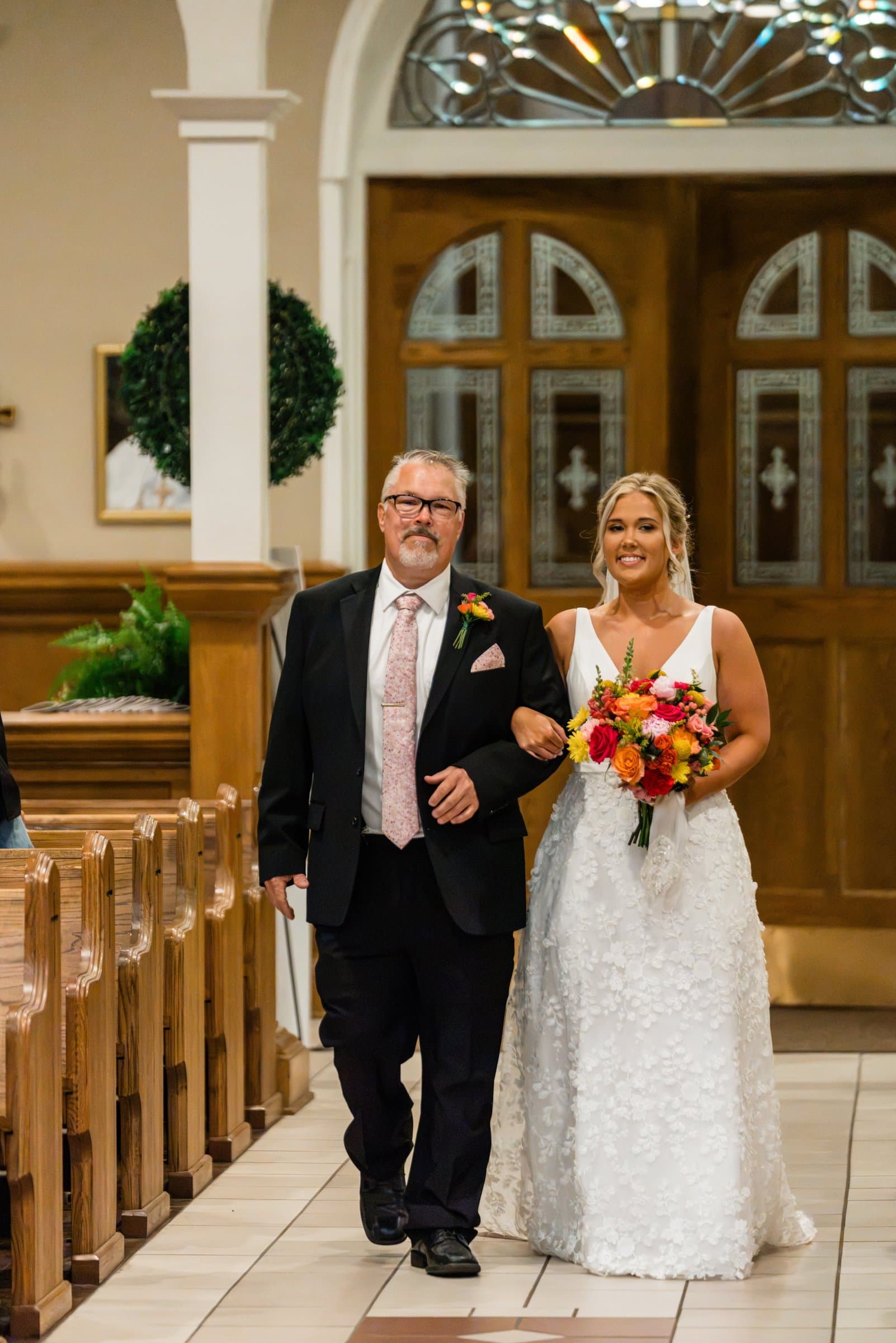 Father walking the bride down the aisle of a church past wooden pews