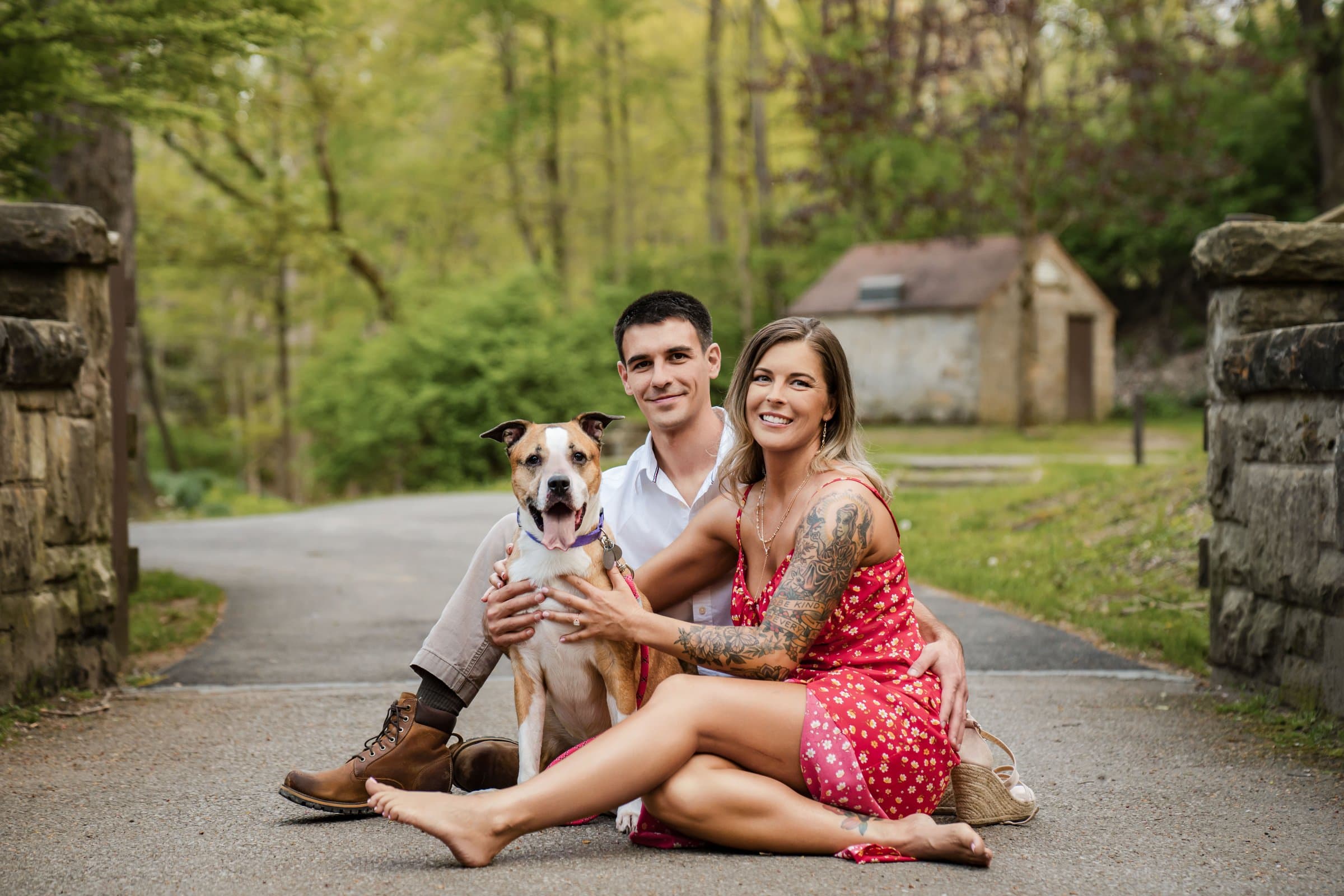 Engaged couple sitting on a park path between stone pillars with their tan and white dog