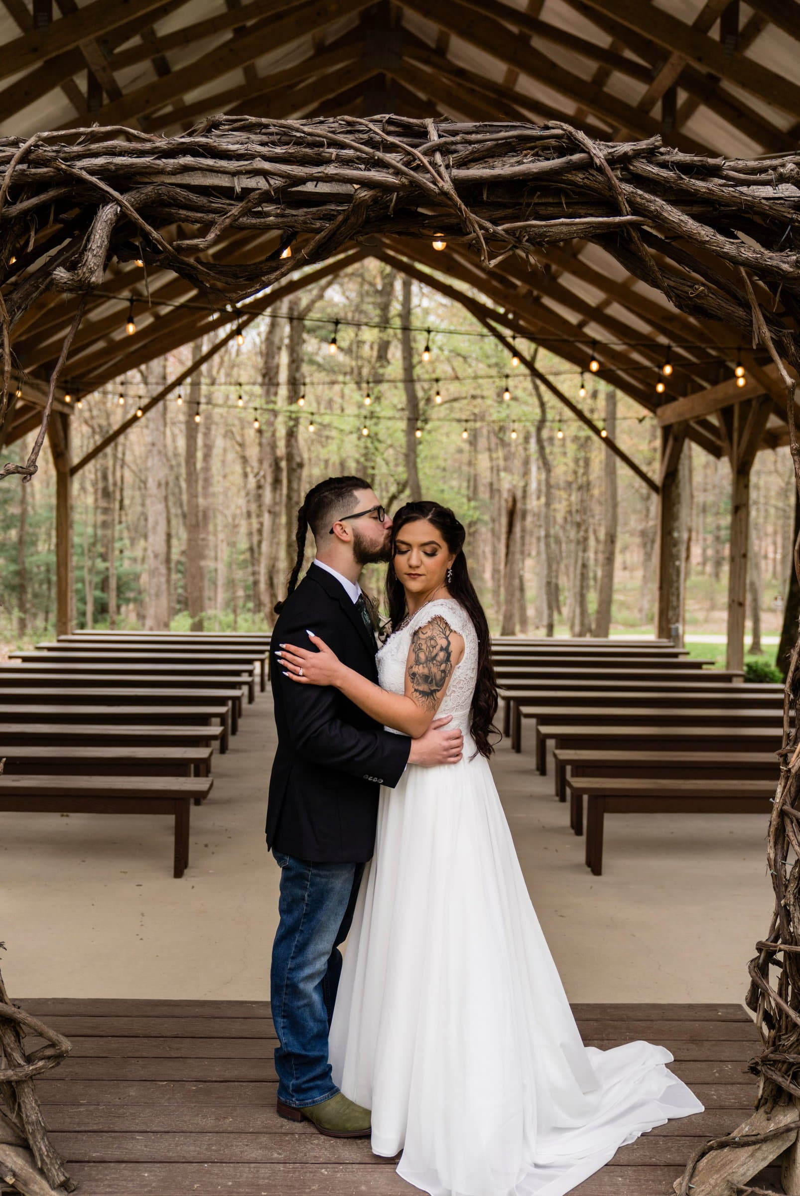 A bride and groom embracing at the end of the aisle in an open-sided wooden wedding chapel, with wooden pews on both sides and bare woods behind.