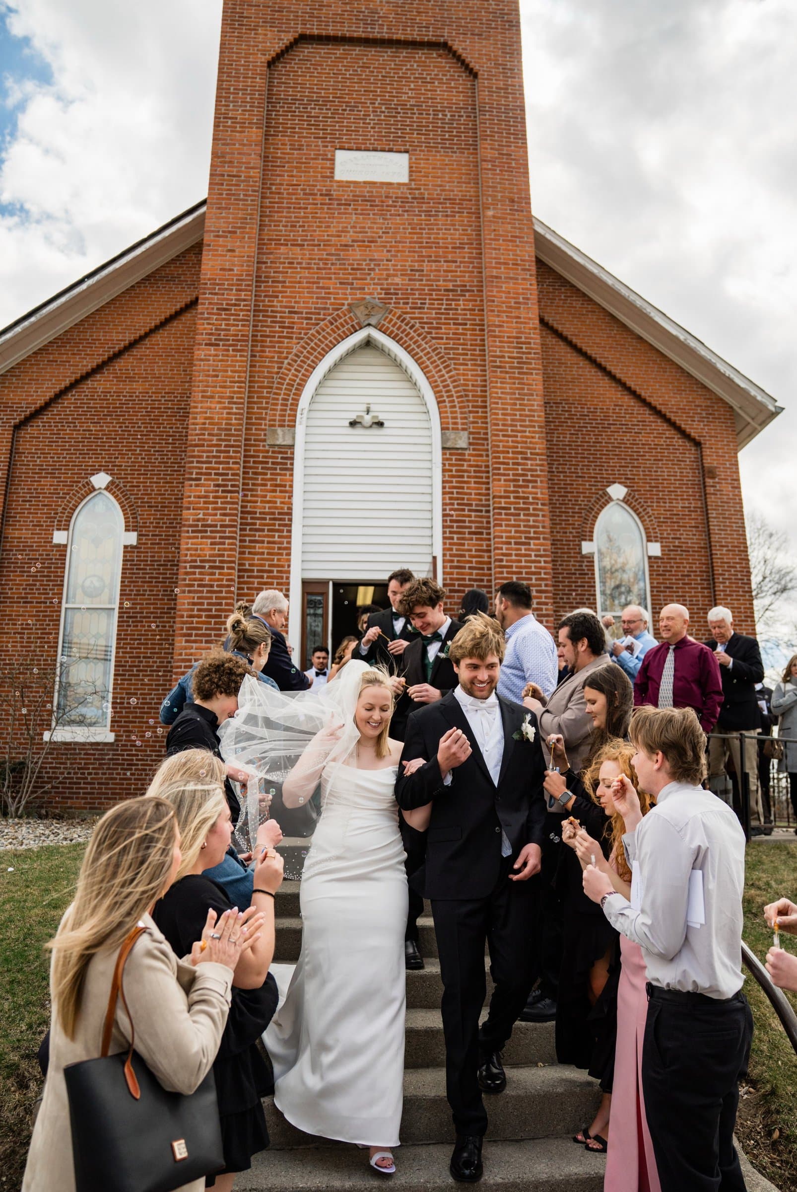 A bride and groom come down the front steps of a red brick church with an arched white door while guests line the stairs ringing bells